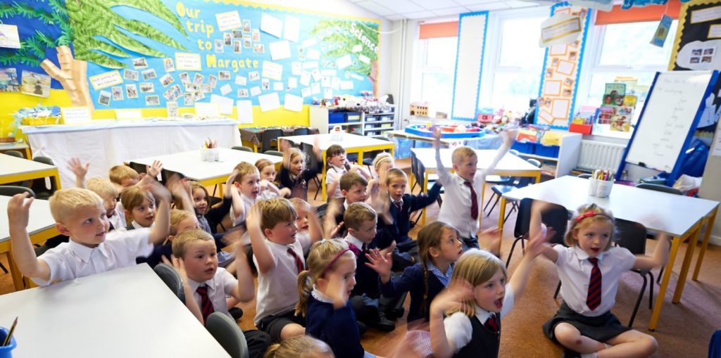 Children in classroom sitting on floor with their hands up