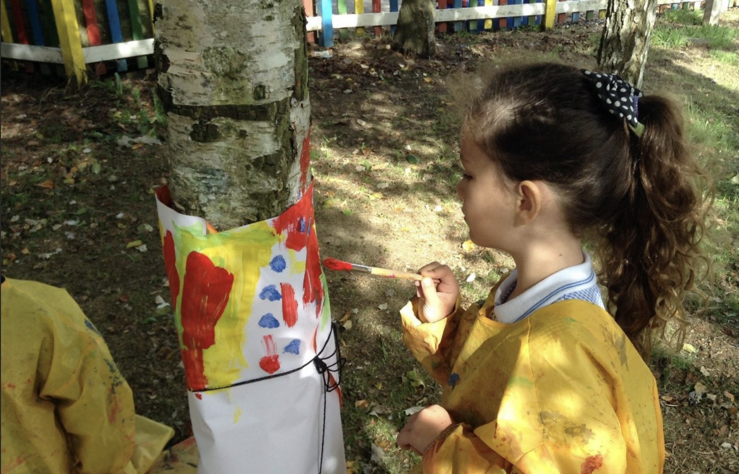 A young girl is seen wearing an art apron and painting on a sheet of paper which is tied to a tree trunk outdoors.